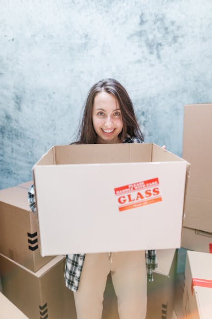 A person with long brown hair smiling and standing indoors against a plain, light-colored wall, holding an empty cardboard box labeled 'FRAGILE GLASS' in red lettering. The individual is dressed in casual clothing, including a plaid shirt and light-colored pants. Behind them are additional cardboard boxes of various sizes, some sealed and others open, positioned on the floor and leaning against the wall, indicating a home relocation or packing process. The scene appears to be part of a furniture transport or house removal operation, demonstrating careful packing and handling of fragile items, and is associated with professional removals services such as those offered by Man and Van Battersea specializing in house removals and moving logistics.