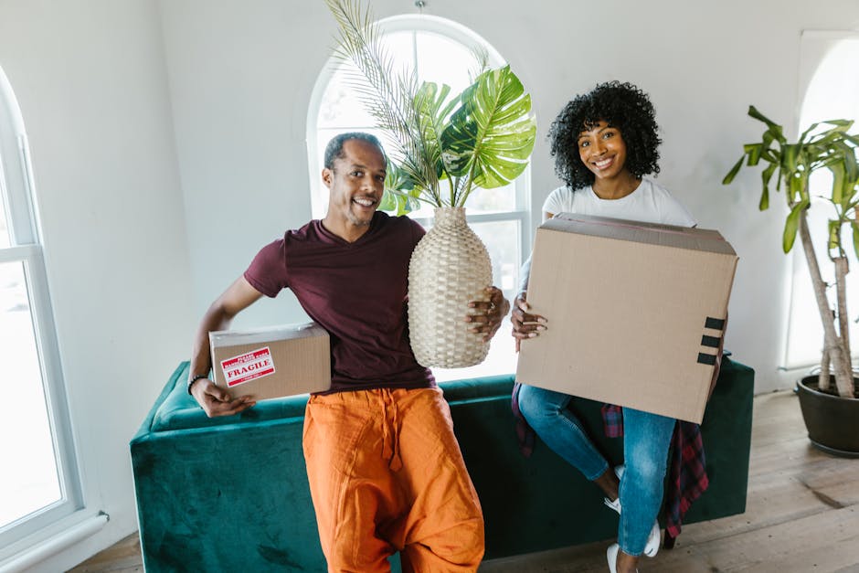 A man and a woman inside a bright, modern living room during a home relocation process, both smiling and holding cardboard boxes filled with household items. The man, dressed in a maroon t-shirt and orange trousers, is holding a small parcel with a 'Fragile' label in one hand and a large, textured ceramic vase with green monstera leaves in the other. The woman, wearing a white t-shirt and blue jeans, is carrying a medium-sized box. They are positioned in front of a green sofa, next to a large window with white trims that allows natural light to illuminate the space. Behind them, there is a tall indoor plant in a black pot. This scene captures the packing and loading process typical of furniture transport and home removal services, with visible packing materials and the casual setting of a residential interior, reflecting the kind of service provided by Man and Van Battersea during house removals in SW11.