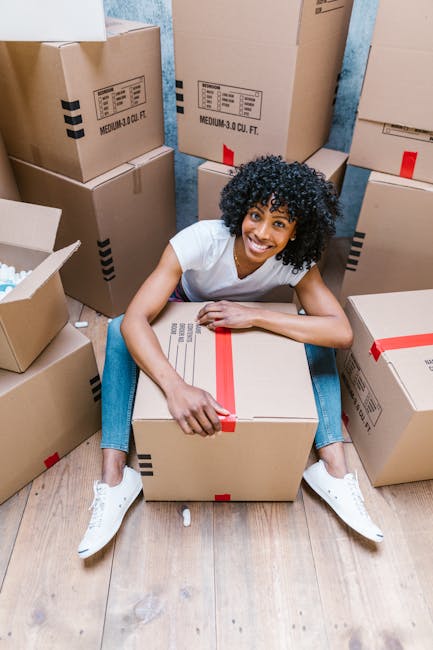 A person with long brown hair smiling and standing indoors against a plain, light-colored wall, holding an empty cardboard box labeled 'FRAGILE GLASS' in red lettering. The individual is dressed in casual clothing, including a plaid shirt and light-colored pants. Behind them are additional cardboard boxes of various sizes, some sealed and others open, positioned on the floor and leaning against the wall, indicating a home relocation or packing process. The scene appears to be part of a furniture transport or house removal operation, demonstrating careful packing and handling of fragile items, and is associated with professional removals services such as those offered by Man and Van Battersea specializing in house removals and moving logistics.