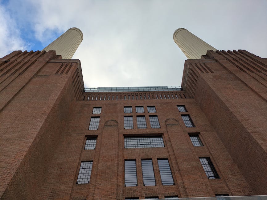 A view of the Battersea Power Station and adjacent modern glass-office building seen from across the River Thames on a cloudy day. The power station, constructed of red brick, features four tall white